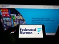 Person holding mobile phone with logo of American investment manager Federated Hermes Inc. on screen in front of web page.