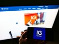 Person holding mobile phone with logo of British financial services company IG Group Holdings Plc. on screen with web page.