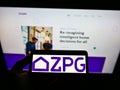 Person holding mobile phone with logo of British real estate company ZPG Ltd on screen in front of business web page.