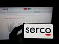 Person holding mobile phone with logo of British service company Serco Group plc on screen in front of business web page.