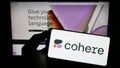 Person holding mobile phone with logo of Canadian AI company Cohere Inc. on screen in front of business web page.