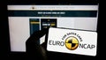 Person holding mobile phone with logo of car safety programme Euro NCAP on screen in front of web page.