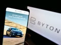 Person holding mobile phone with logo of Chinese automotive brand Byton on screen in front of business web page.