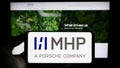Person holding mobile phone with logo of company MHP Management- und IT-Beratung GmbH on screen in front of web page.