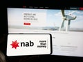 Person holding mobile phone with logo of company National Australia Bank Limited (NAB) on screen in front of web page.
