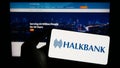 Person holding mobile phone with logo of company Turkiye Halk Bankasi A.S. (Halkbank) on screen in front of web page.