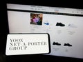 Person holding mobile phone with logo of company YOOX Net-a-Porter Group S.p.A. (YNAP) on screen in front of web page.