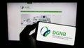 Person holding mobile phone with logo of Deutsche Gesellschaft fÃ¼r Nachhaltiges Bauen (DGNB) on screen with webpage.