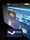 Person holding mobile phone with logo of distribution platform Steam operated by Valve Corp on screen in front of web page.