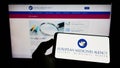 Person holding mobile phone with logo of EU agency European Medicines Agency (EMA) on screen in front of web page.