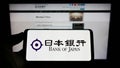 Person holding mobile phone with logo of financial institution Bank of Japan (BOJ) on screen in front of web page.