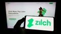 Person holding mobile phone with logo of fintech Zilch Technology Limited (PayZilch) on screen in front of web page.