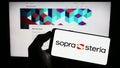 Person holding mobile phone with logo of French company Sopra Steria Group SA on screen in front of business web page.