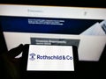 Person holding mobile phone with logo of French financial services company Rothschild Co. SCA on screen in front of web page.