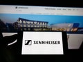 Person holding mobile phone with logo of German audio company Sennheiser electronic GmbH Co KG on screen in front of webpage.