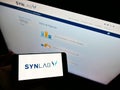 Person holding mobile phone with logo of German medical diagnostics company Synlab Group on screen in front of web page.