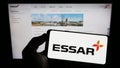 Person holding mobile phone with logo of Indian conglomerate Essar Group on screen in front of business web page.