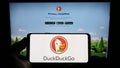 Person holding mobile phone with logo of Internet search engine DuckDuckGo (DDG) on screen in front of company web page.