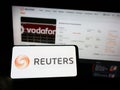 Person holding mobile phone with logo of news agency Reuters on screen in front of company web page.