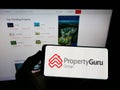 Person holding mobile phone with logo of online property company PropertyGuru Pte. Ltd. on screen in front of web page.
