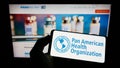 Person holding mobile phone with logo of Pan American Health Organization (PAHO) on screen in front of web page. Royalty Free Stock Photo