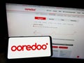 Person holding mobile phone with logo of Qatari telecommunications company Ooredoo QSC on screen in front of web page.