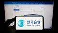 Person holding mobile phone with logo of South Korean central bank Bank of Korea (BOK) on screen in front of web page.