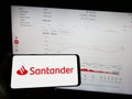 Person holding mobile phone with logo of Spanish banking company Banco Santander S.A. on screen in front of web page.