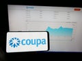 Person holding mobile phone with logo of spend management company Coupa Software Inc. on screen in front of web page.