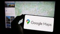 Person holding mobile phone with logo of web mapping platform Google Maps on screen in front of web page.