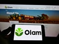 Person holding smartphone with business logo of agribusiness company Olam International Ltd on screen in front of website.