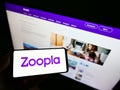 Person holding smartphone with business logo of British real estate platform Zoopla Limited on screen in front of webpage.