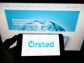 Person holding smartphone with business logo of Danish energy company ÃËrsted AS on screen in front of web page.