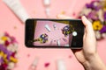 A person is holding a smartphone in his hand and taking a perfect flat lay photo with skincare products and colorful flowers on a