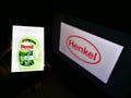 Person holding smartphone with laundry detergent product Persil on display marketed by company Henkel with logo in background. Royalty Free Stock Photo