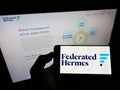 Person holding smartphone with logo of American investment manager Federated Hermes Inc. on screen in front of webpage.