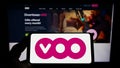 Person holding smartphone with logo of Belgian telecommunications company Voo S.A. on screen in front of website.