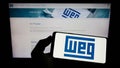 Person holding smartphone with logo of Brazilian company WEG Equipamentos Eletricos S.A. on screen in front of website.