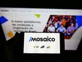 Person holding smartphone with logo of Brazilian e-commerce platform Mosaico Tecnologia on screen in front of website.