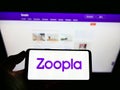 Person holding smartphone with logo of British real estate platform Zoopla Limited on screen in front of website.