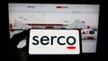 Person holding smartphone with logo of British service company Serco Group plc on screen in front of website.