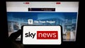Person holding smartphone with logo of British television channel Sky News on screen in front of website.