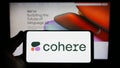 Person holding smartphone with logo of Canadian AI company Cohere Inc. on screen in front of website.