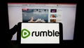 Person holding smartphone with logo of Canadian video platform company Rumble Inc. on screen in front of website. Royalty Free Stock Photo