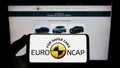 Person holding smartphone with logo of car safety programme Euro NCAP on screen in front of website.