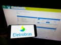 Person holding smartphone with logo of Centrais ElÃÂ©tricas Brasileiras S.A. (Eletrobras) on screen in front of website. Royalty Free Stock Photo