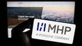 Person holding smartphone with logo of company MHP Management- und IT-Beratung GmbH on screen in front of website.