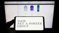 Person holding smartphone with logo of company YOOX Net-a-Porter Group S.p.A. (YNAP) on screen in front of website.