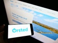 Person holding smartphone with logo of Danish energy company ÃËrsted AS on screen in front of website.