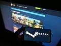 Person holding smartphone with logo of distribution platform Steam operated by Valve on screen in front of website.